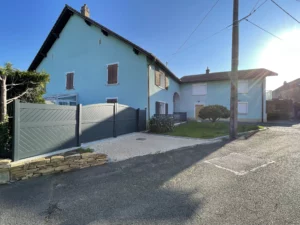 Portail battant en aluminium gris, installé devant une maison bleue avec une allée en gravier, baignée par la lumière du soleil