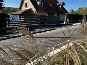 Portail et clôture en aluminium noir, installés devant une maison avec une allée asphaltée, vue à travers des herbes sèches