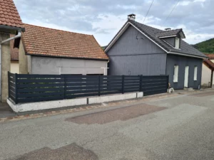 Portail manuel en aluminium gris, installé devant une maison avec des façades en bois et en béton